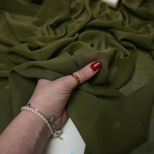 K&ouml;nnte beinhalten: Olivgr&uuml;ner Stoff mit flie&szlig;ender, transparenter Textur. Eine Hand mit einem goldenen Ring und einem Armband h&auml;lt den Stoff und zeigt seinen Fall. Eine Garnrolle befindet sich im Hintergrund. Der Stoff wird wahrscheinlich f&uuml;r Bekleidung oder Bastelprojekte verwendet.