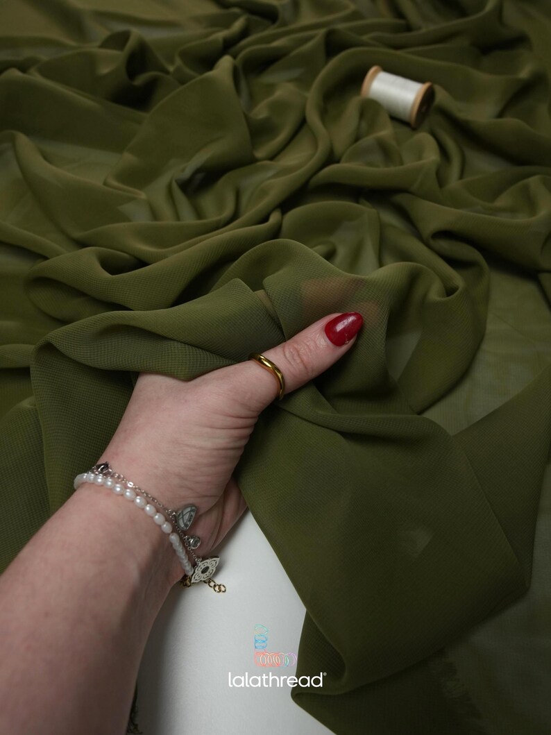 K&ouml;nnte beinhalten: Olivgr&uuml;ner Stoff mit flie&szlig;ender, transparenter Textur. Eine Hand mit einem goldenen Ring und einem Armband h&auml;lt den Stoff und zeigt seinen Fall. Eine Garnrolle befindet sich im Hintergrund. Der Stoff wird wahrscheinlich f&uuml;r Bekleidung oder Bastelprojekte verwendet.