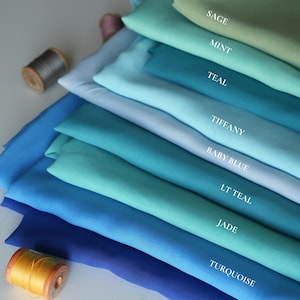 K&ouml;nnte beinhalten: Stoffmuster in verschiedenen Blau- und Gr&uuml;nt&ouml;nen, von Salbei bis K&ouml;nigsblau. Die Farben sind beschriftet, mit sichtbaren Garnrollen. Das Bild zeigt eine Auswahl an Textilfarben.