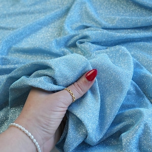 May include: Light blue fabric with a shimmering, glittery texture. The fabric is draped, creating soft folds and shadows. The material appears to be a lightweight textile, possibly for apparel or decorative use.