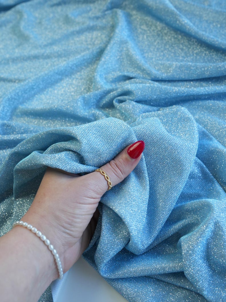May include: Light blue fabric with a shimmering, glittery texture. The fabric is draped, creating soft folds and shadows. The material appears to be a lightweight textile, possibly for apparel or decorative use.