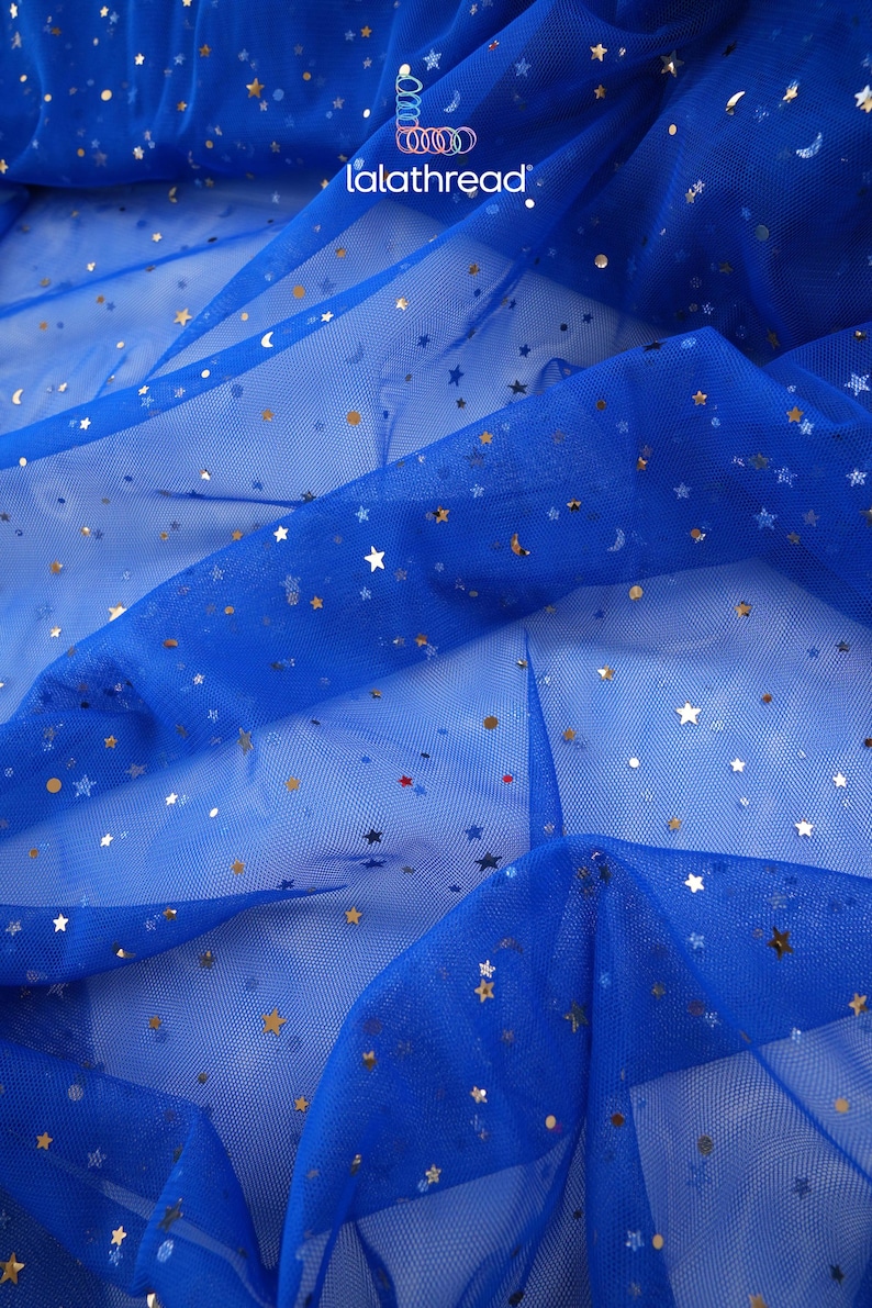 May include: Royal blue tulle fabric scattered with gold stars, moons, and small dots. The fabric is semi-sheer and has a delicate, flowing appearance. The brand name "lalathread" is visible in the upper right corner. This fabric is suitable for crafting and apparel.