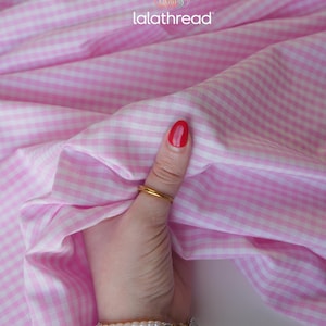 Puede incluir: Tela de cuadros vichy rosa y blanco con el texto "lalathread". La tela est&aacute; sujeta por una mano con un anillo dorado y una pulsera. El esmalte de u&ntilde;as es rojo.