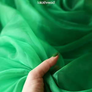May include: Close-up of a hand holding a swatch of vibrant green fabric. The fabric is sheer and appears to be silk or a similar lightweight material. The logo "lalathread" is visible in the upper right corner.