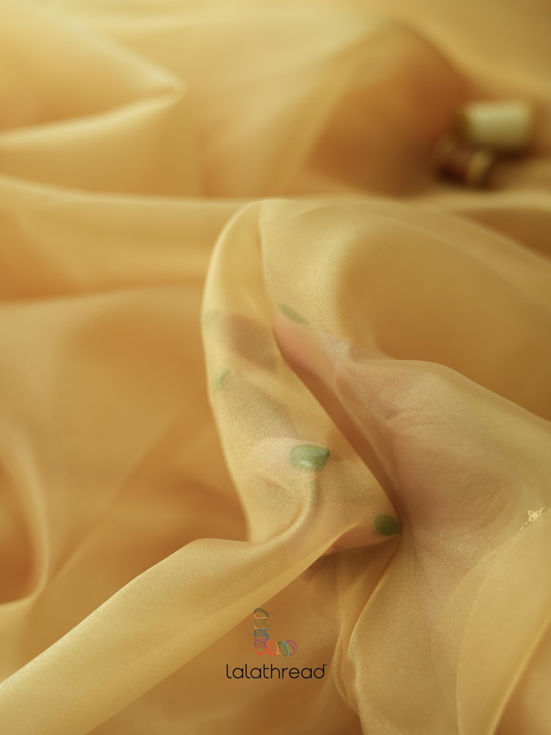 May include: A close-up of a sheer, light gold fabric with a subtle shimmer. The fabric is draped and folded, creating soft, flowing lines. The fabric is translucent, allowing light to pass through it.