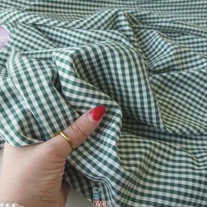 May include: Close-up of green and white gingham fabric. The fabric is folded and draped, showing the pattern's detail. The fabric is being held by a hand with a gold ring and red nail polish. The brand name "lalathread" is visible.