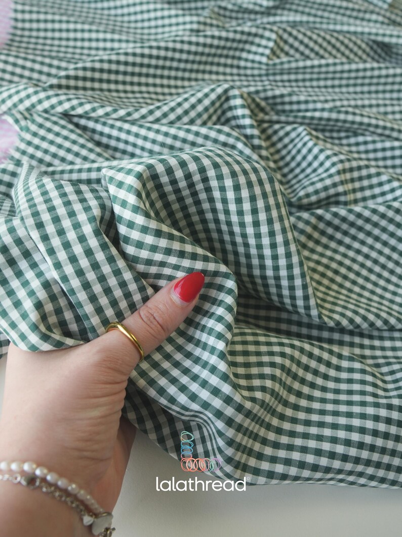 May include: Close-up of green and white gingham fabric. The fabric is folded and draped, showing the pattern's detail. The fabric is being held by a hand with a gold ring and red nail polish. The brand name "lalathread" is visible.