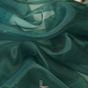 May include: Close-up of flowing, sheer teal fabric with a subtle sheen. The fabric is draped, creating soft folds and shadows. The brand name "lalathread" is visible in the lower right corner. Two spools of thread are in the background.