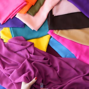 K&ouml;nnte beinhalten: Eine lebendige Auswahl an farbenfrohen Stoffmustern in Pink-, Lila-, Orange-, Gr&uuml;n-, Gelb- und Blaut&ouml;nen. Eine Hand mit rotem Nagellack h&auml;lt ein St&uuml;ck lila Stoff. Das Logo "lalathread" ist sichtbar.