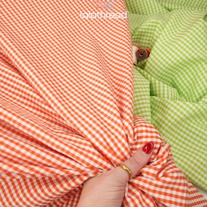 May include: Close-up of orange and white gingham fabric, held by a hand with a gold ring and red nail polish. A second fabric in green and white gingham is visible in the background. The watermark "lalathread" is visible.