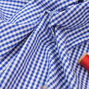 May include: Close-up of blue and white gingham fabric with soft folds. A spool of red thread sits in the corner. The fabric is likely cotton or a similar material, suitable for clothing or home decor projects.
