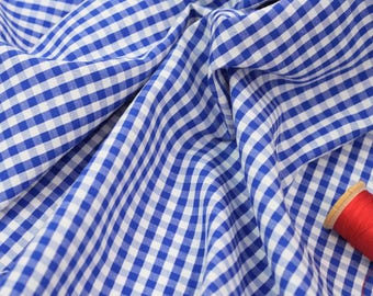 May include: Close-up of blue and white gingham fabric with soft folds. A spool of red thread sits in the corner. The fabric is likely cotton or a similar material, suitable for clothing or home decor projects.