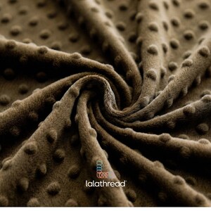 May include: A close-up shot of a brown fabric with a textured, raised dot pattern. The material appears soft and plush, with a swirling design. The logo "lalathread" is visible. This fabric is suitable for blankets, throws, and other textile projects.
