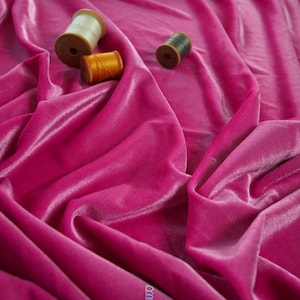 May include: A close-up shot of vibrant fuchsia velvet fabric with soft folds. Three spools of thread in cream, orange, and dark green are scattered across the fabric. The fabric has a luxurious sheen, suggesting a high-quality textile.