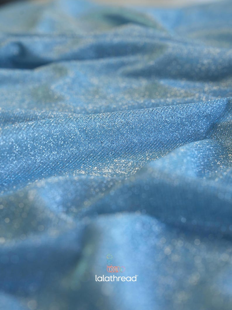 May include: Close-up of a light blue fabric with a shimmering, glittery texture. The fabric is draped, creating soft folds and shadows. The word "lalathread" is visible in the bottom right corner.