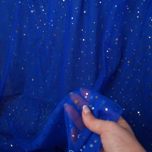 May include: Royal blue tulle fabric with gold and silver star and crescent moon embellishments. The fabric is semi-sheer and has a delicate texture. The image shows a close-up of the fabric, with a hand holding a portion of it. The brand name "lalathread" is visible.