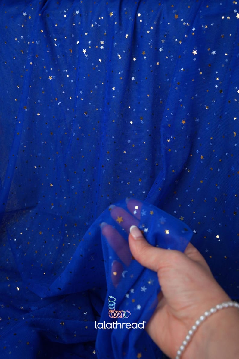 May include: Royal blue tulle fabric with gold and silver star and crescent moon embellishments. The fabric is semi-sheer and has a delicate texture. The image shows a close-up of the fabric, with a hand holding a portion of it. The brand name "lalathread" is visible.