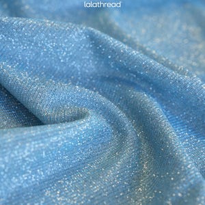 May include: Close-up of a light blue fabric with a shimmering, glittery texture. The fabric is draped, creating soft folds and shadows. The text "lalathread" is visible in the upper left corner.