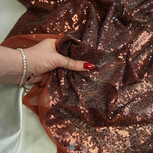 May include: Close-up of a shimmering brown sequin fabric. The fabric is held up, revealing a sheer, rust-colored backing. The sequins catch the light, creating a sparkling effect. The fabric is likely used for apparel or decorative purposes.