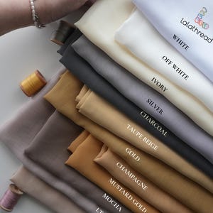 K&ouml;nnte beinhalten: Eine Auswahl an Stoffmustern in verschiedenen Farben, darunter Wei&szlig;, Elfenbein, Silber und Gold. Die Farben sind beschriftet. Eine Hand h&auml;lt die Muster. Auch Garnrollen sind zu sehen.