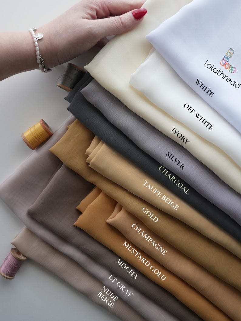 K&ouml;nnte beinhalten: Eine Auswahl an Stoffmustern in verschiedenen Farben, darunter Wei&szlig;, Elfenbein, Silber und Gold. Die Farben sind beschriftet. Eine Hand h&auml;lt die Muster. Auch Garnrollen sind zu sehen.