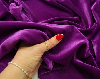 May include: A close-up of a vibrant purple velvet fabric. The fabric is draped and folded, creating shadows and highlights that emphasize its texture. The fabric is being held by a hand with a gold ring and a pearl bracelet.