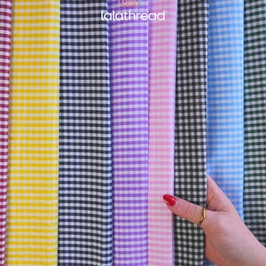 May include: A variety of gingham fabric in multiple colors, including yellow, black, purple, pink, blue, and green. The fabric is arranged vertically, with a hand holding the black and white checkered fabric. The text "lalathread" is visible.