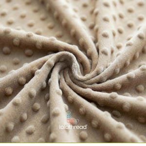 May include: A close-up shot of a beige fabric with a textured, dotted pattern. The fabric has a soft, plush appearance with a swirling design. The logo "lalathread" is visible. This fabric is suitable for blankets, clothing, or other textile projects.