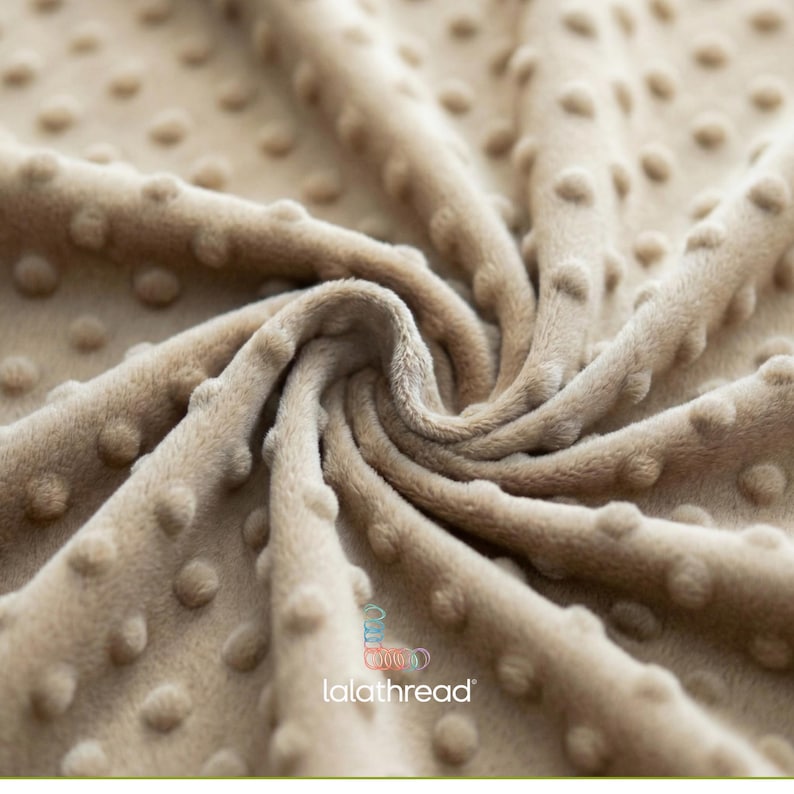 May include: A close-up shot of a beige fabric with a textured, dotted pattern. The fabric has a soft, plush appearance with a swirling design. The logo "lalathread" is visible. This fabric is suitable for blankets, clothing, or other textile projects.