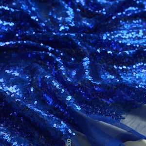 May include: Close-up of a vibrant blue sequin fabric. The fabric is heavily textured with countless reflective sequins, creating a shimmering effect. The material is draped, showing folds and shadows. The brand name "lalathread" is visible.