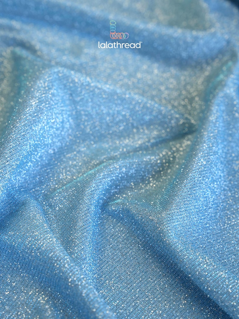 May include: Close-up of a shimmering, light blue fabric with a textured surface. The material has a subtle sparkle, catching the light and creating a luminous effect. The fabric is likely used for apparel or decorative purposes.