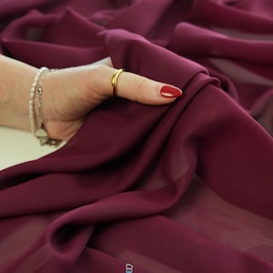 May include: Close-up of a deep burgundy fabric with soft folds. The fabric is held by a hand with a gold ring and red nail polish. A spool of thread is visible in the background. The logo "lalathread" is in the bottom right corner.