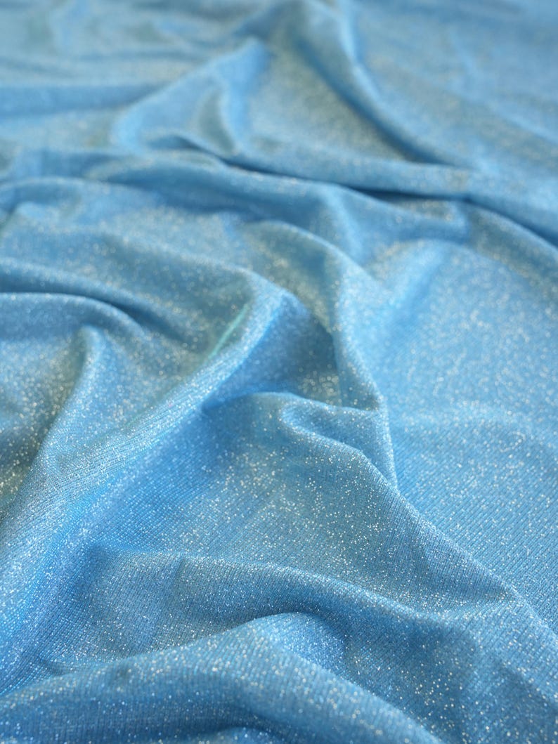 May include: Close-up of a light blue fabric with a subtle sparkle. The material is draped, creating soft folds and shadows. The fabric has a shimmering quality, suggesting it might be used for apparel or decorative purposes.