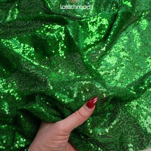May include: Bright green sequin fabric with a shiny, reflective surface. The fabric is densely covered in small, round sequins, creating a shimmering effect. The fabric is draped, showing its texture and flexibility. The brand name "lalathread" is visible in the upper right corner.