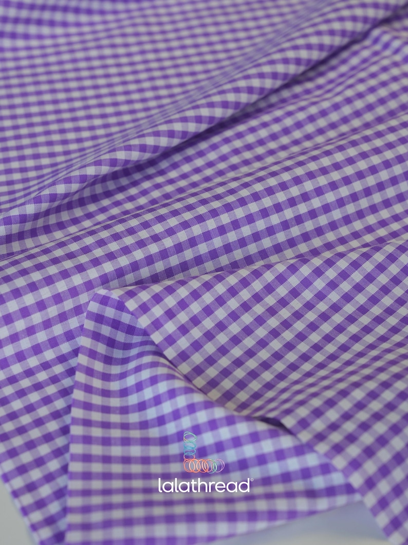 Puede incluir: Primer plano de una tela con estampado de cuadros vichy morados y blancos. La tela est&aacute; doblada, mostrando la textura y la ca&iacute;da del material. La imagen est&aacute; bien iluminada, resaltando el color y el patr&oacute;n. La tela es adecuada para proyectos de costura o manualidades.