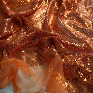 May include: Close-up of shimmering orange sequin fabric. The fabric is draped, creating folds and shadows that highlight the texture and sparkle of the sequins. A sheer orange tulle is visible in the foreground. The logo "lalathread" is in the bottom right corner.