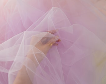 May include: A close-up of layers of pastel-colored tulle fabric. The fabric is a mix of pink, lavender, and peach hues. The word "lalathread" is printed on the fabric in a colorful, stylized font. A hand is holding the fabric.