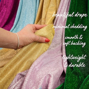 May include: A hand holds a swatch of gold fabric from a collection of shimmering textiles. The image features various colors, including pink, green, blue, and gold. Text on the image reads "Designed to shed less - shimmer more" and highlights features like "beautiful drape" and "lightweight & durable."