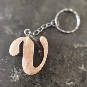 May include: A glittery, rose-gold letter "V" keychain. The resin-cast letter is attached to a silver chain and keyring. The keychain is a personalized accessory, suitable for bags, keys, or as a decorative item.