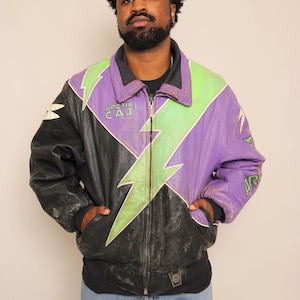 Peut inclure: Veste en cuir noire, violette et verte avec un motif d'éclair. La veste porte le texte "ARCTIC CAT" sur la poitrine et les manches. Portée sur un t-shirt noir et un jean bleu.