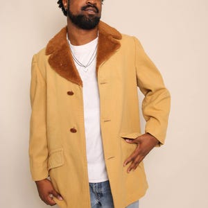 Op de afbeelding: Mosterdgele jas met een bruine imitatiebontkraag. De jas heeft een knoopsluiting, twee voorzakken en een klassiek, getailleerd ontwerp. Het model draagt een wit t-shirt en een blauwe jeans.