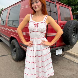 May include: A white midi dress featuring a subtle polka dot pattern and thin red piping accents along the neckline, straps, and waist. The bodice includes a small red bow detail, and the skirt is adorned with horizontal red stripes.