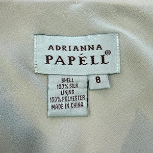 K&ouml;nnte beinhalten: Ein wei&szlig;es Etikett mit braunem Text, auf dem "Adrianna Papell" und "Shell 100% Silk Lining 100% Polyester Made in China" steht. Das Etikett hat auch ein kleineres wei&szlig;es Etikett mit der Nummer "8" darauf.