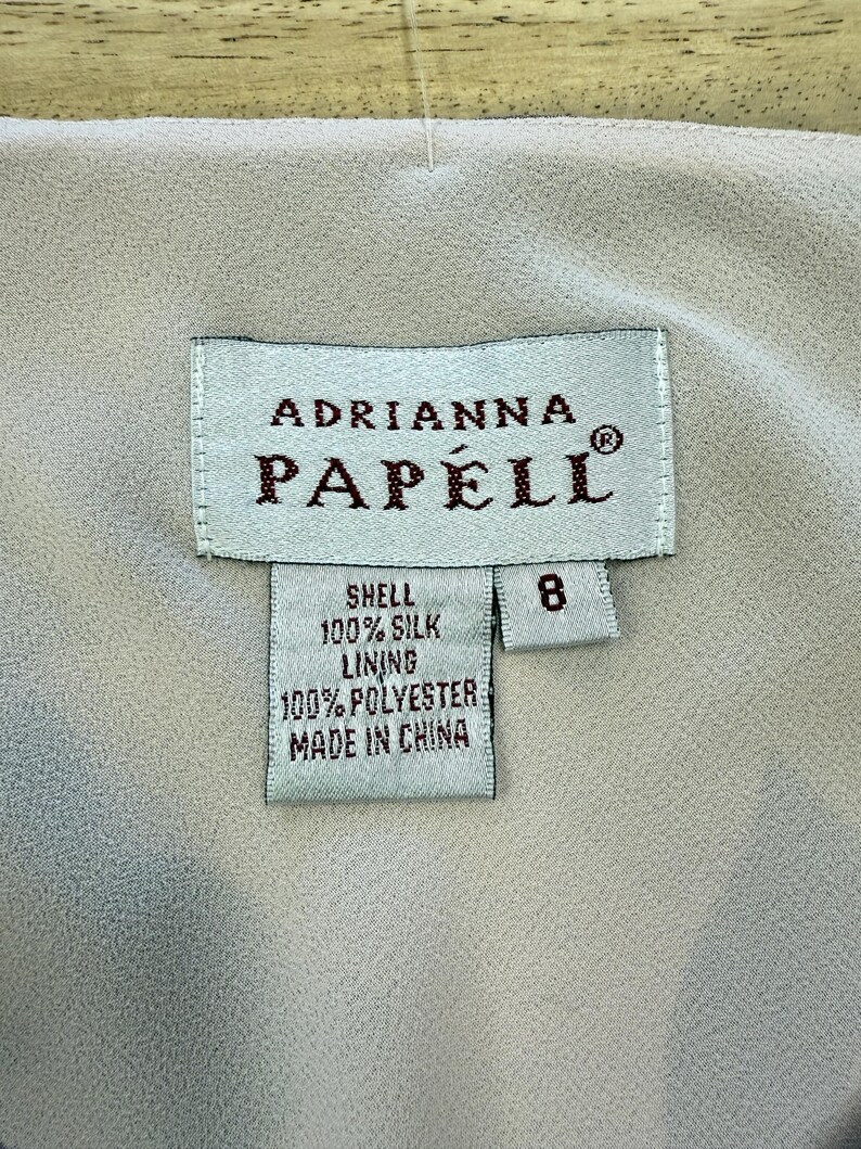 K&ouml;nnte beinhalten: Ein wei&szlig;es Etikett mit braunem Text, auf dem "Adrianna Papell" und "Shell 100% Silk Lining 100% Polyester Made in China" steht. Das Etikett hat auch ein kleineres wei&szlig;es Etikett mit der Nummer "8" darauf.