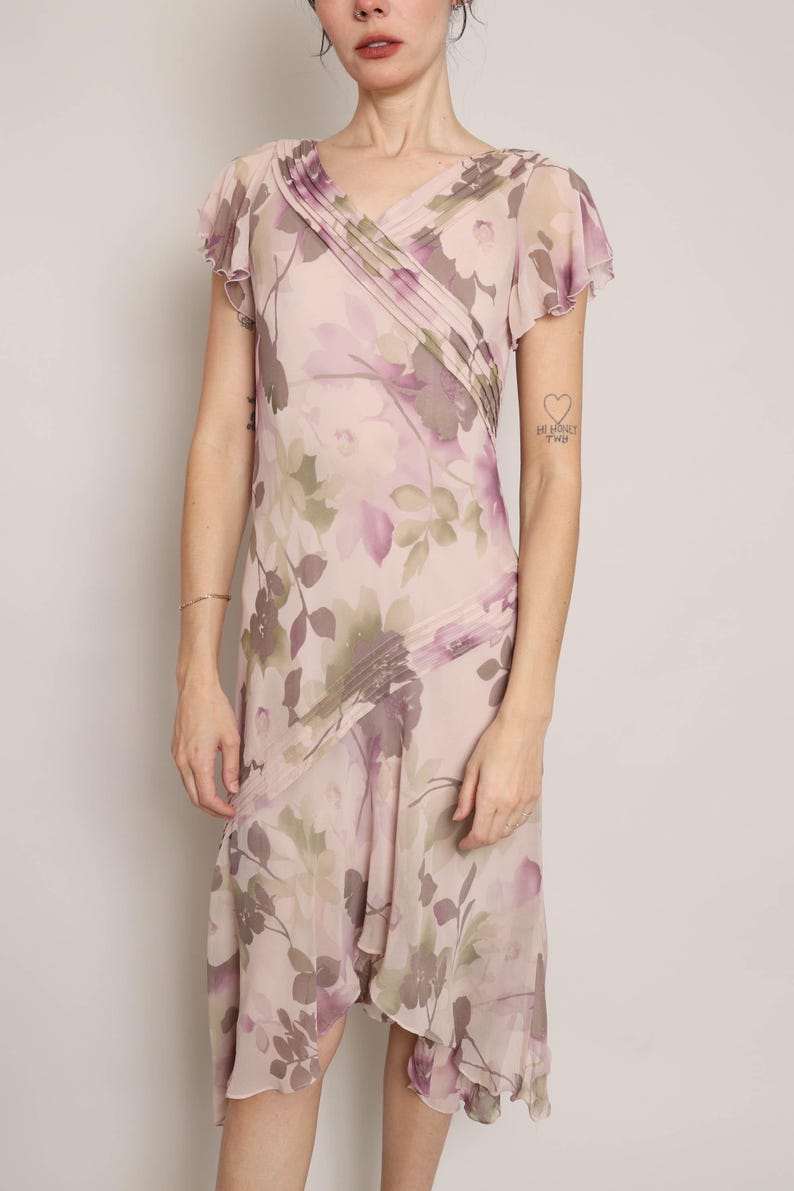 K&ouml;nnte beinhalten: Ein rosa-violettes Kleid mit floralem Muster, V-Ausschnitt und kurzen &Auml;rmeln. Das Kleid hat einen flie&szlig;enden, asymmetrischen Saum.