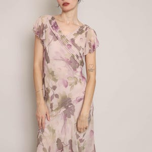 K&ouml;nnte beinhalten: Ein rosafarbenes und violettes Midi-Kleid mit Blumenmuster, V-Ausschnitt und kurzen &Auml;rmeln. Das Kleid hat einen R&uuml;schen-Saum und einen figurbetonten Oberteil. Das Model tr&auml;gt ein silbernes Armband am linken Handgelenk und hat ein kleines Tattoo auf ihrem linken Arm, auf dem "Ich liebe dich" steht.