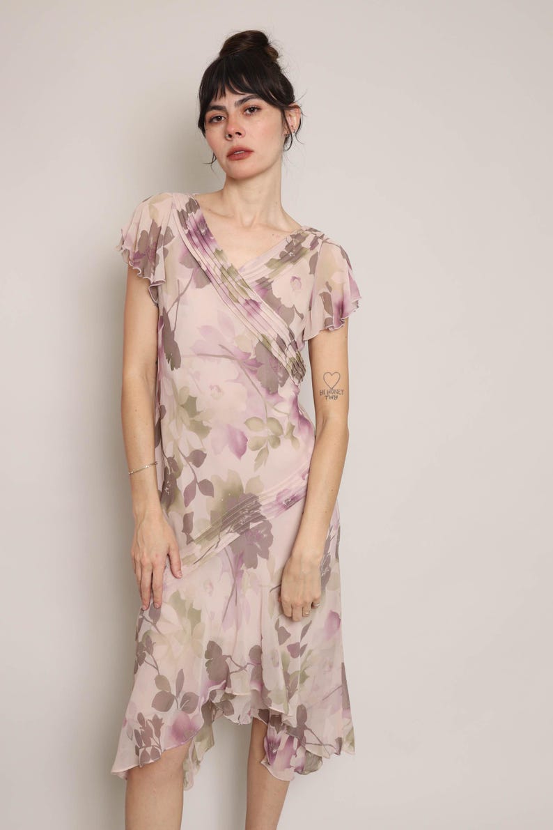 K&ouml;nnte beinhalten: Ein rosafarbenes und violettes Midi-Kleid mit Blumenmuster, V-Ausschnitt und kurzen &Auml;rmeln. Das Kleid hat einen R&uuml;schen-Saum und einen figurbetonten Oberteil. Das Model tr&auml;gt ein silbernes Armband am linken Handgelenk und hat ein kleines Tattoo auf ihrem linken Arm, auf dem "Ich liebe dich" steht.