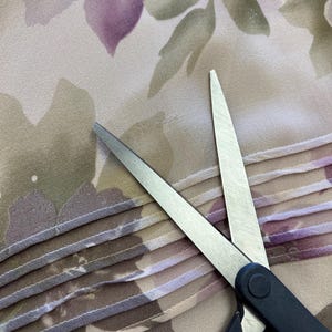 K&ouml;nnte beinhalten: Eine silberne Schere mit schwarzen Griffen schneidet ein St&uuml;ck beigefarbenen Stoffes mit einem Blumenmuster. Der Stoff hat einen gefalteten Rand.