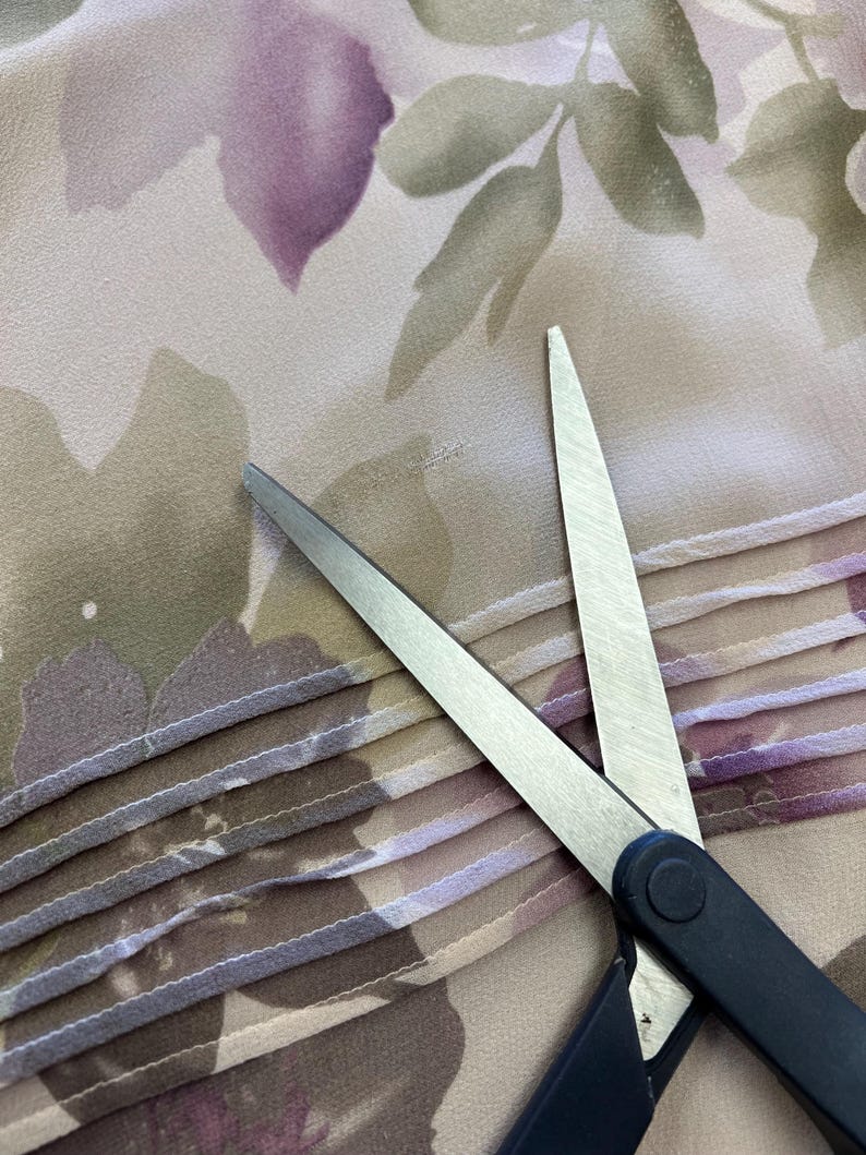 K&ouml;nnte beinhalten: Eine silberne Schere mit schwarzen Griffen schneidet ein St&uuml;ck beigefarbenen Stoffes mit einem Blumenmuster. Der Stoff hat einen gefalteten Rand.