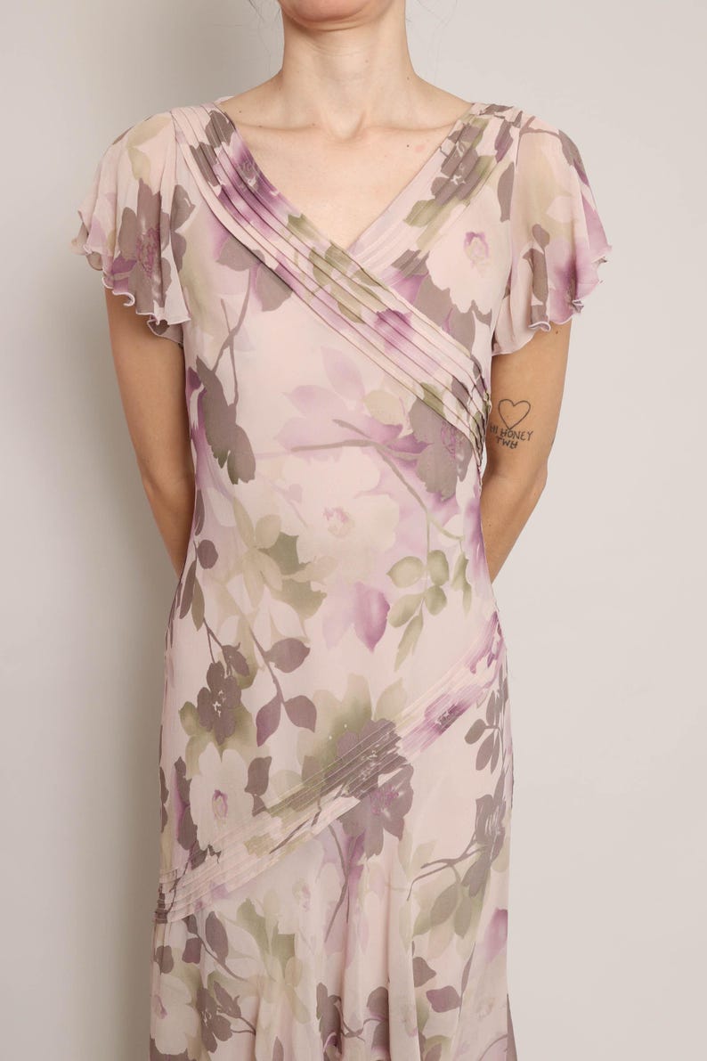 K&ouml;nnte beinhalten: Ein rosa Kleid mit floralem Muster, V-Ausschnitt und kurzen &Auml;rmeln. Das Kleid hat eine Faltendetail am Ausschnitt und einen flie&szlig;enden Rock.